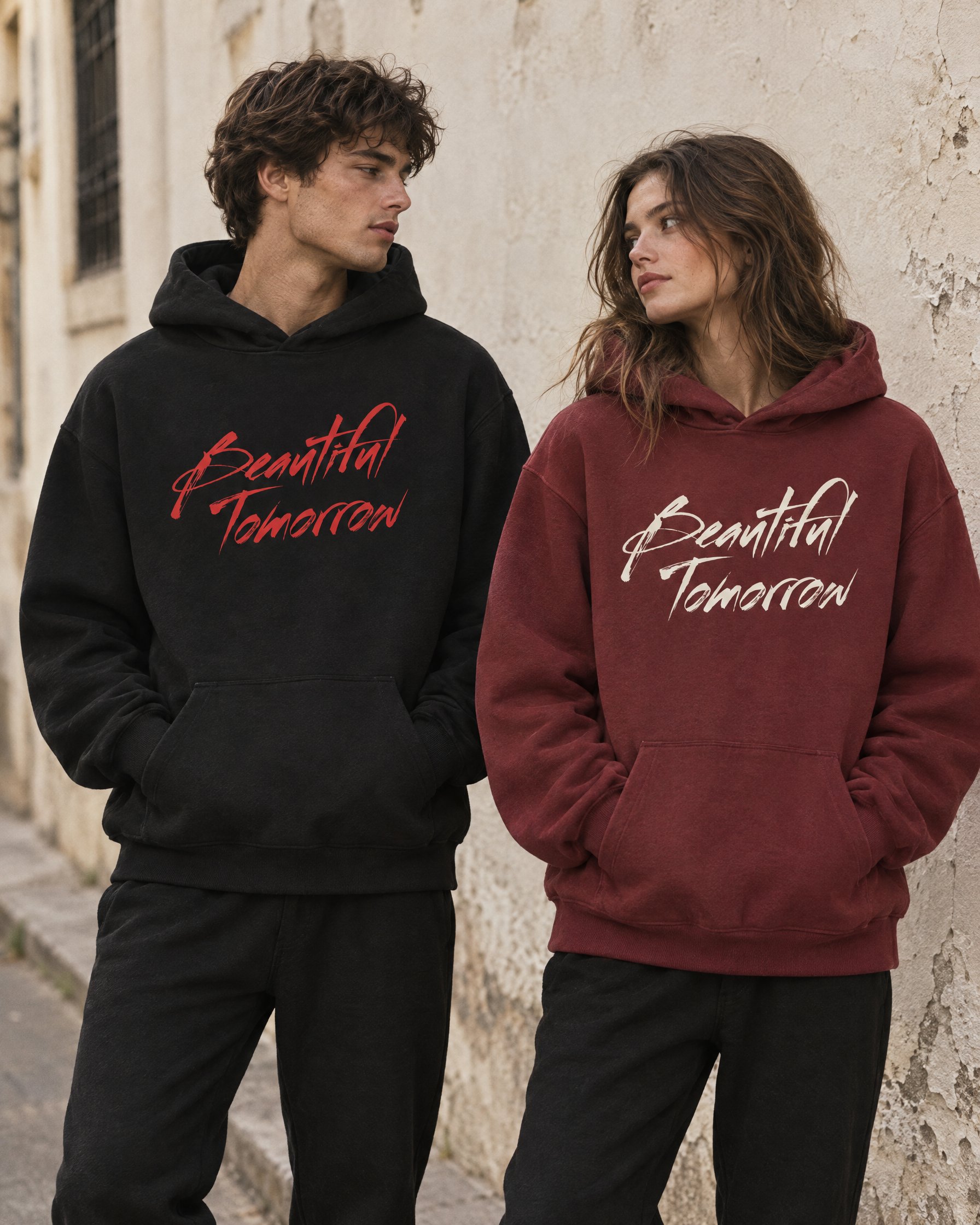 Two models against a plaster wall, one in the black Classic Hoodie with the red brush-script Beautiful Tomorrow logo, one in the red Classic Hoodie with the logo in white