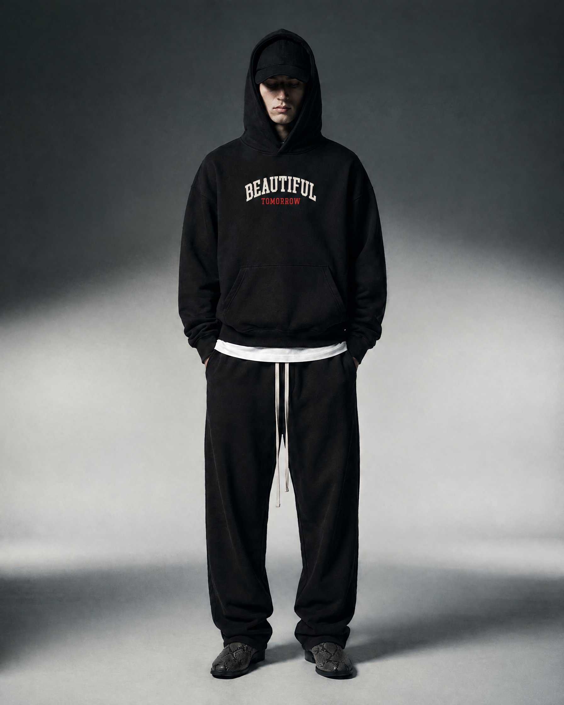 Model in the black Varsity Hoodie with red TOMORROW lettering, hood up over a cap, with wide black sweatpants